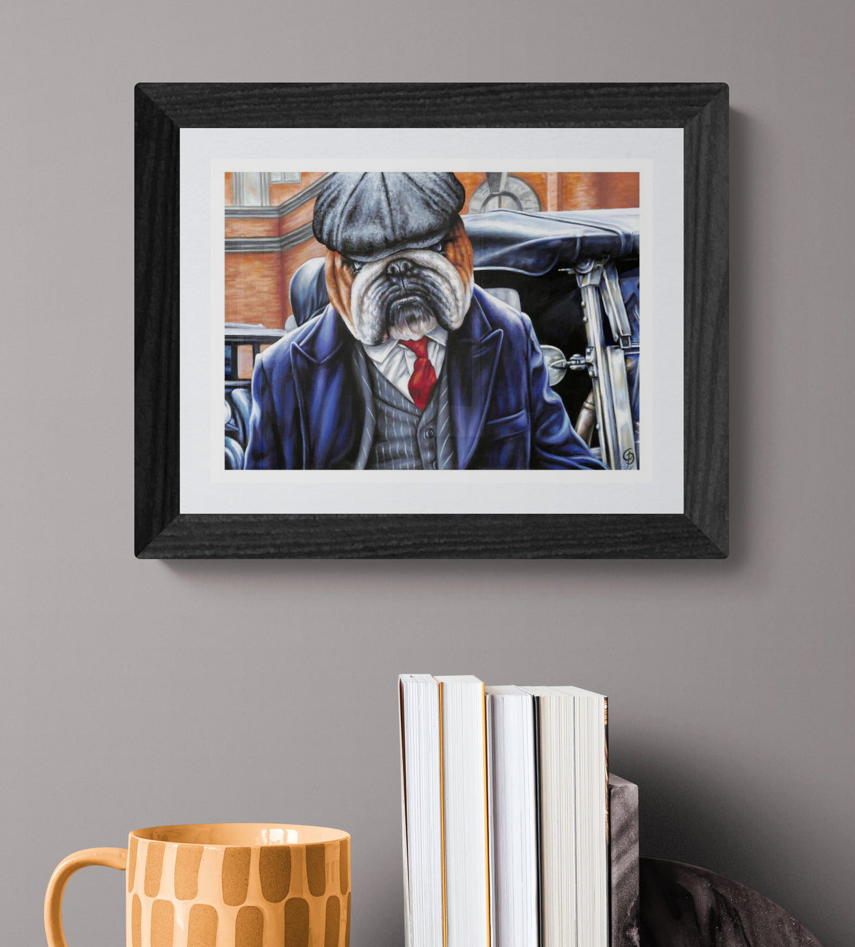 Framed artwork of a bulldog in a suit on a gray wall with books and a mug below.
