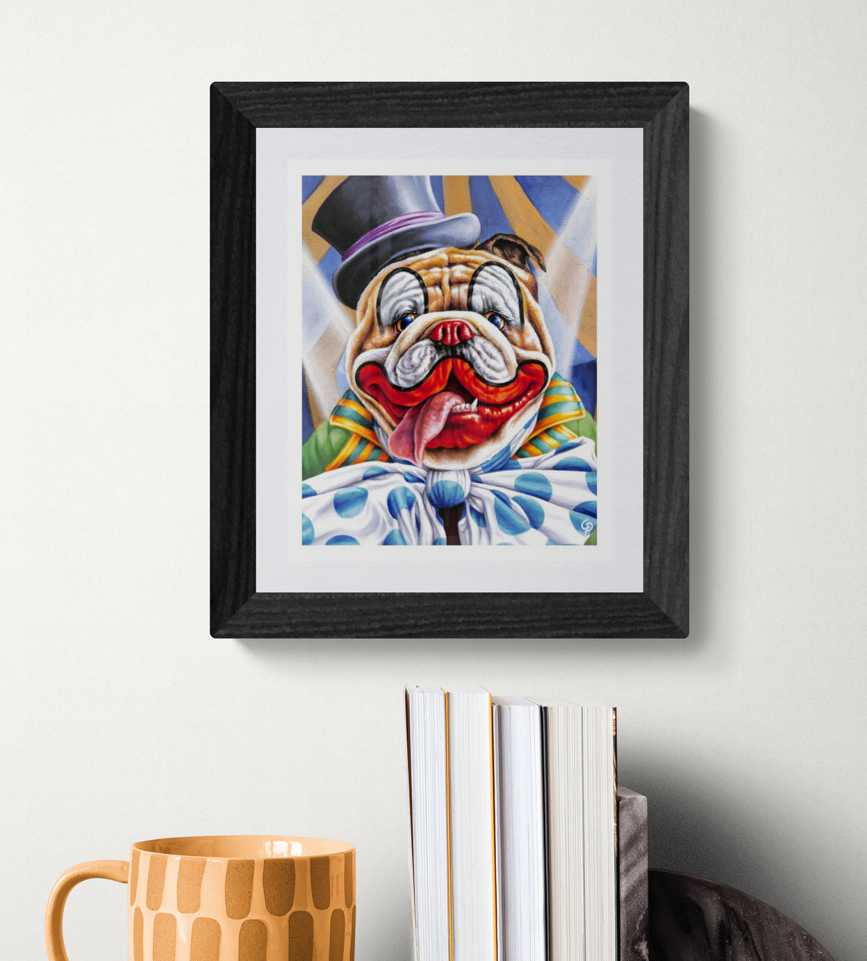 Framed artwork of a bulldog in clown attire on a white wall with books and a mug below.