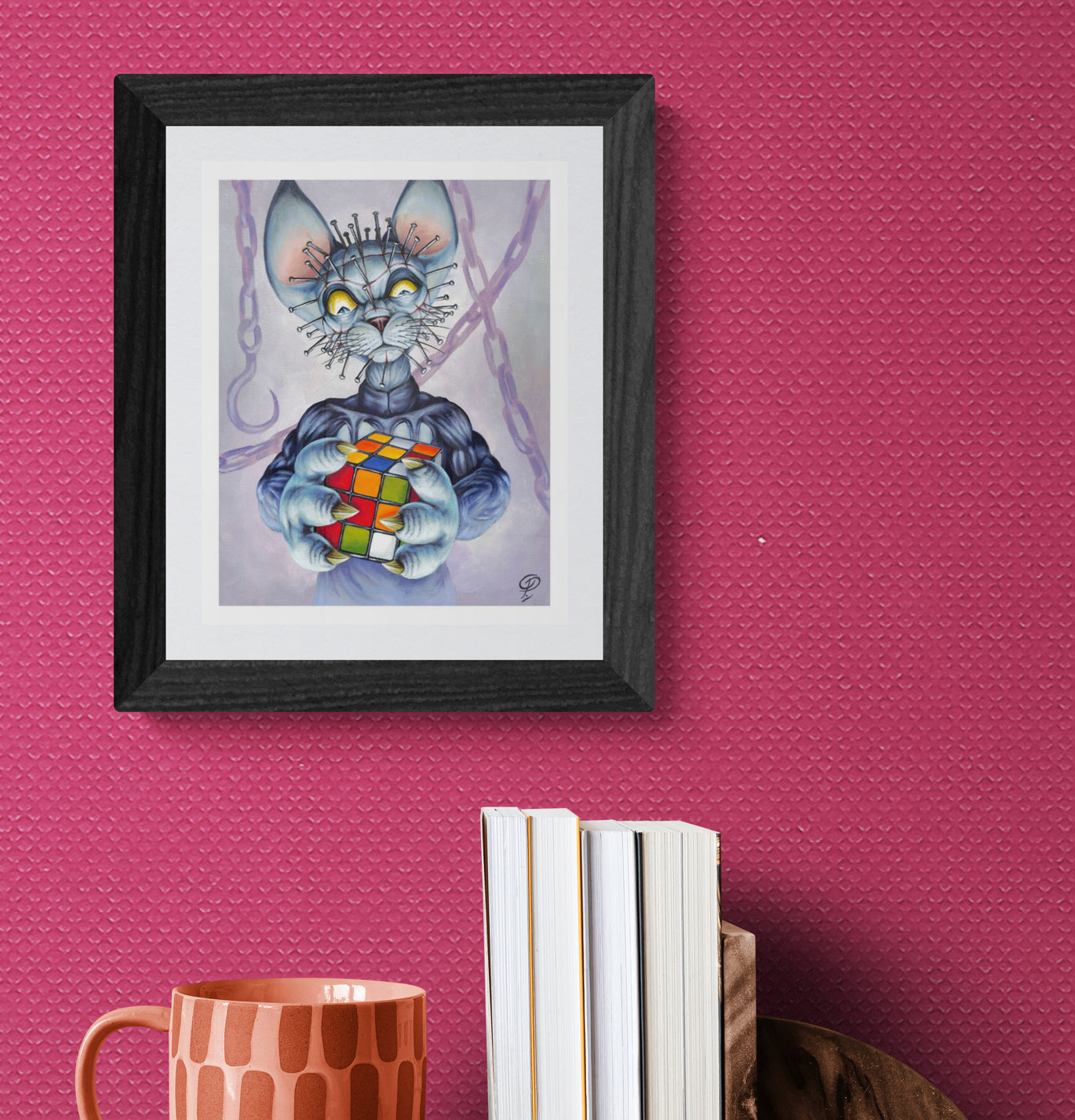 Framed artwork of a cat holding a Rubik's Cube on a pink wall with books and a mug below.