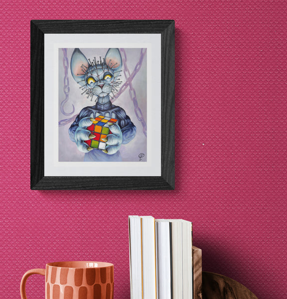 Framed artwork of a cat holding a Rubik's Cube on a pink wall with books and a mug below.