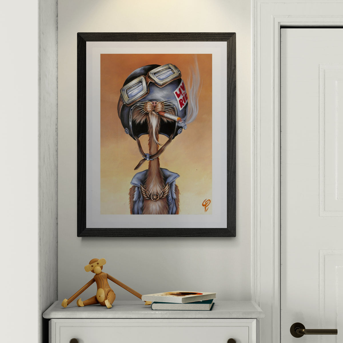 Framed artwork of a cat wearing a helmet on a wall above a white cabinet.