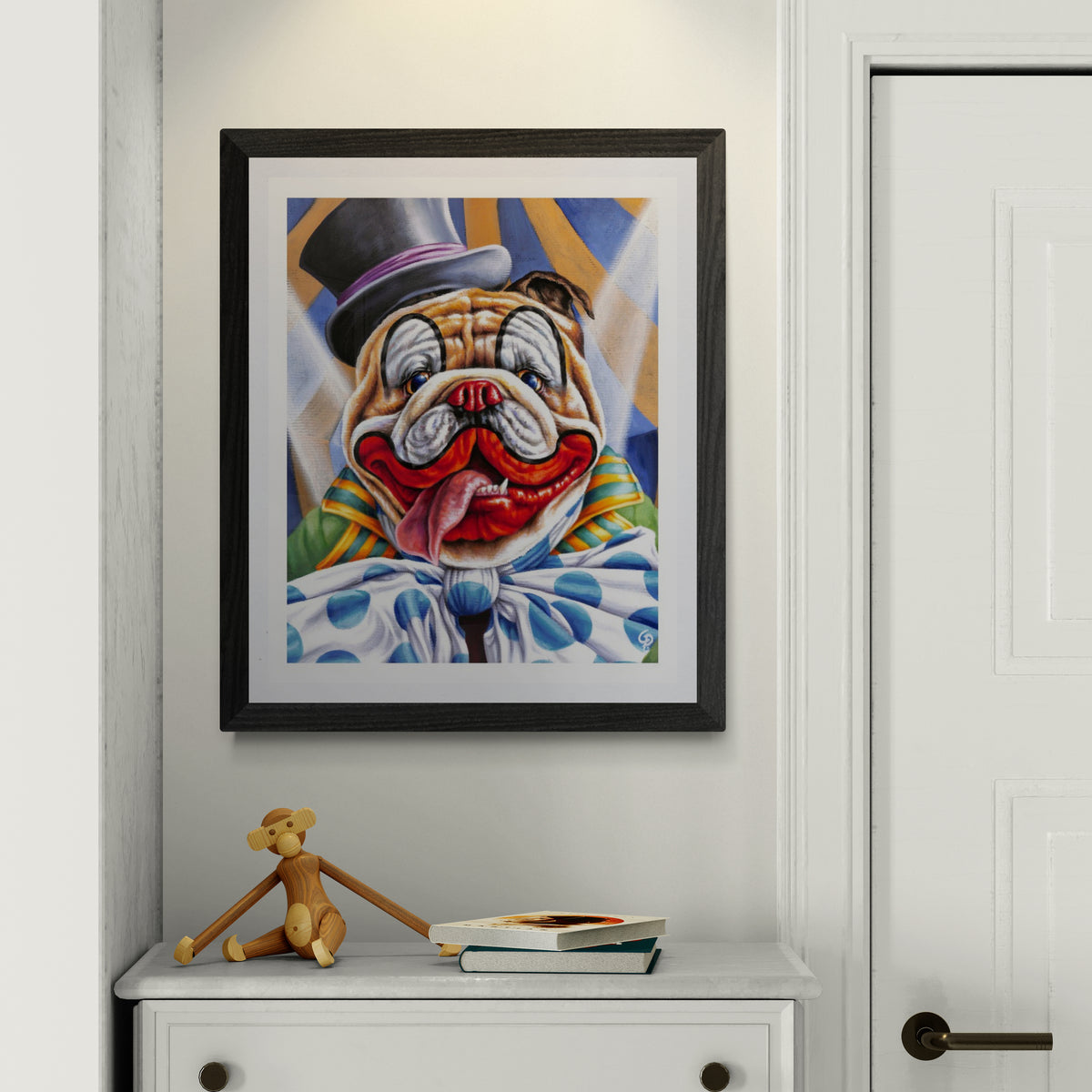 Framed artwork of a clown bulldog on a wall above a white cabinet with a toy and books.